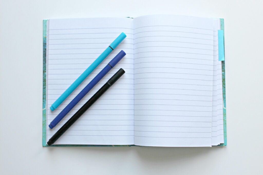 Open lined notebook with colorful pens on a white surface, perfect for note-taking and office tasks.